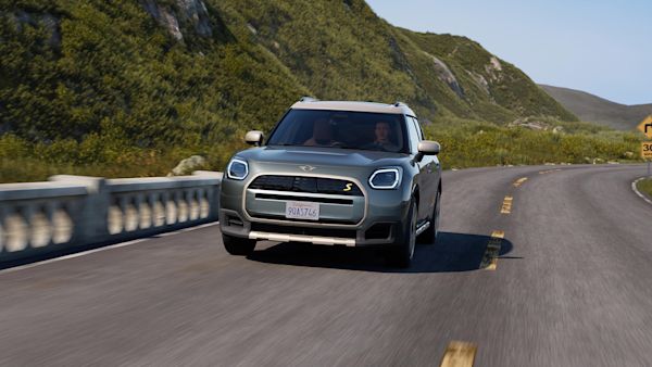 Silver MINI Countryman S driving on a curved mountain road with green hills and a clear blue sky in the background.