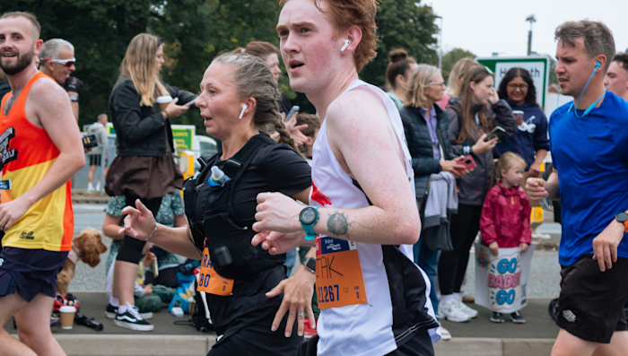 A Shelter runner running alongside other runners in the race. Behind the runners are a group of spectators.