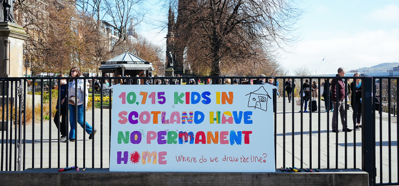 A banner attached to a railing in Princes Street Gardens in Edinburgh. The banner is written in colourful letters and says that 10,715 kids in Scotland have no permanent home.