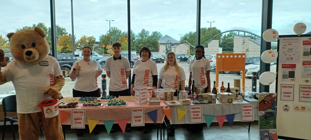 Tables with cakes and prizes. A teddy mascot is in front of the tables wearing a Shelter top ad holding a donations bucket. People are stood behind the table wearing Shelter tops.