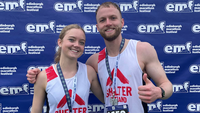 Two smiling runners celebrate after their race, wearing medals and Shelter Scotland running vests. The backdrop has the Edinburgh Marathon Festival logo.