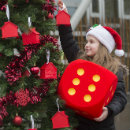 A girl wearing a Santa hat standing next to a Christmas tree and holding a large red dice.