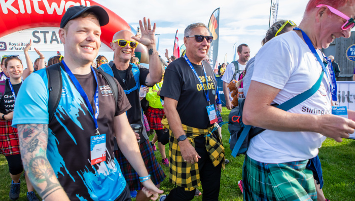 A group of men smile as they start their kiltwalk. 3 of them are wearing sunglasses.