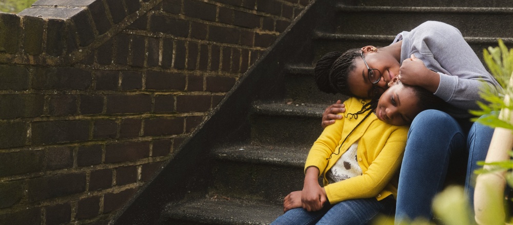 Mother and daughter sit together on the steps of some housing 