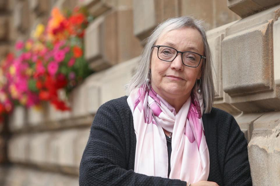 SallyAnn Kelly, chair of Shelter Scotland. She's standing in front of a sandstone wall and a colourful basket of flowers. She's wearing glasses, a back jumper and a pink patterned scarf