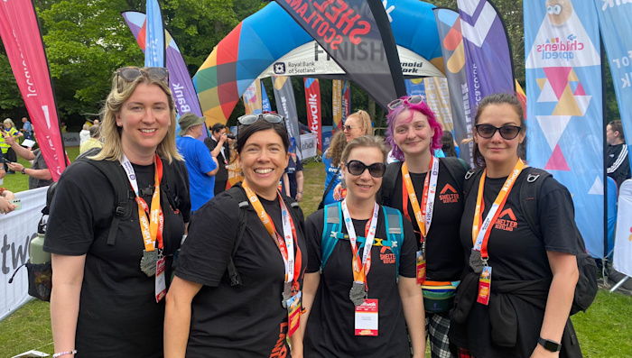 A group of women wearing Shelter Scotland tops and tartan trousers smile at the Kiltwalk finish line.