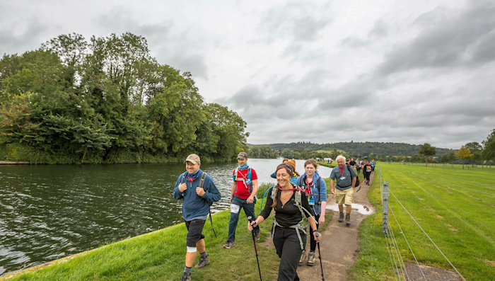 A group of Thames Path Challenge walkers with backpacks and hiking poles. They are hiking along the riverside with green fields and trees in the background.
