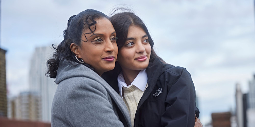 Natalie hugs her daughter Tia, they are both standing on a rooftop looking out over city buildings.