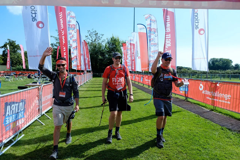 Three walkers wave triumphantly as they cross the finish line of the Lake District Challenge, with colourful banners on either side.
