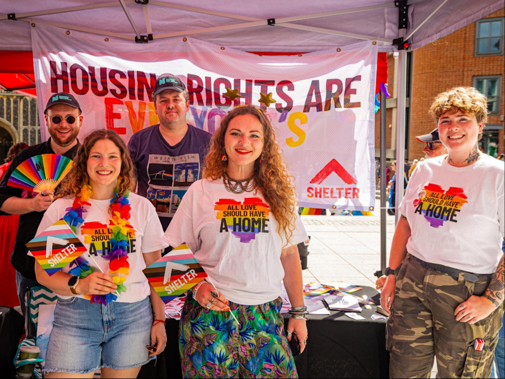 Five young people standing in front of a sign with the Shelter logo and wearing Pride-themed t-shirts.