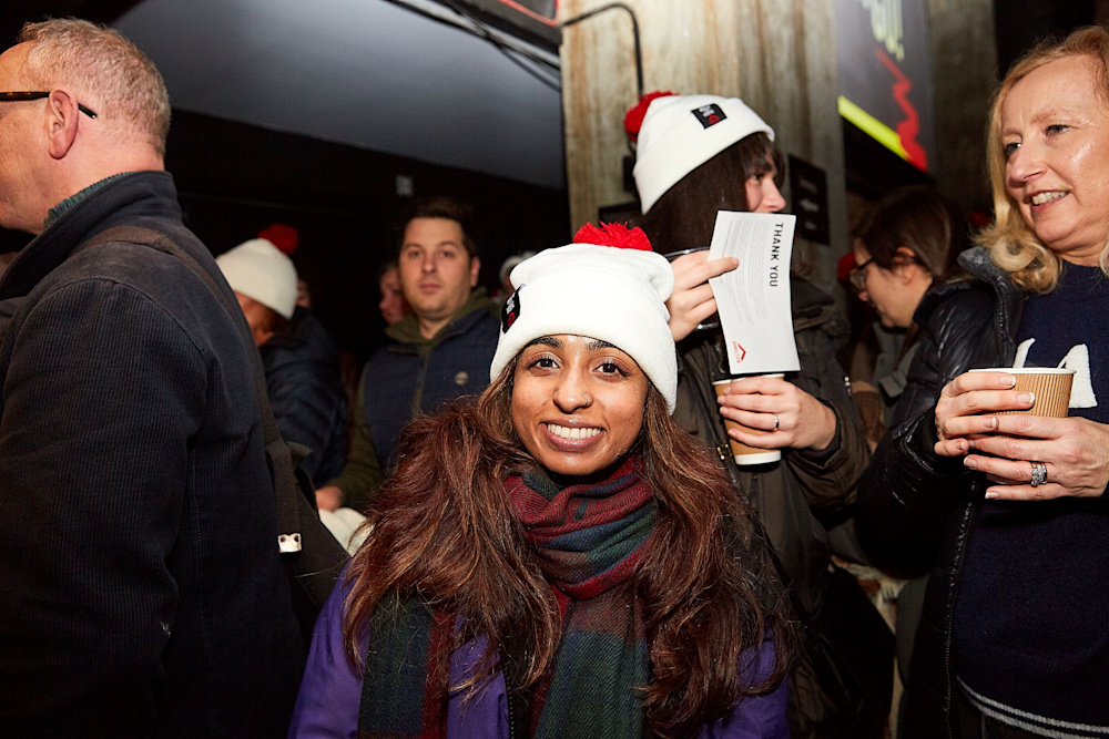 A woman wearing a white and red Walk for Home bobble hat. She is standing in a bustling crowd with people chatting and carrying coffee cups.