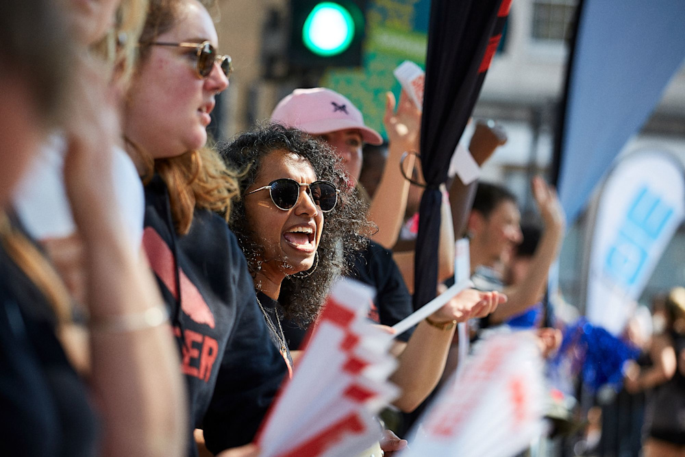 Shelter staff and volunteers at the cheer point of a race, wearing Shelter hoodies and sunglasses. They are clapping and waving to the passing runners.