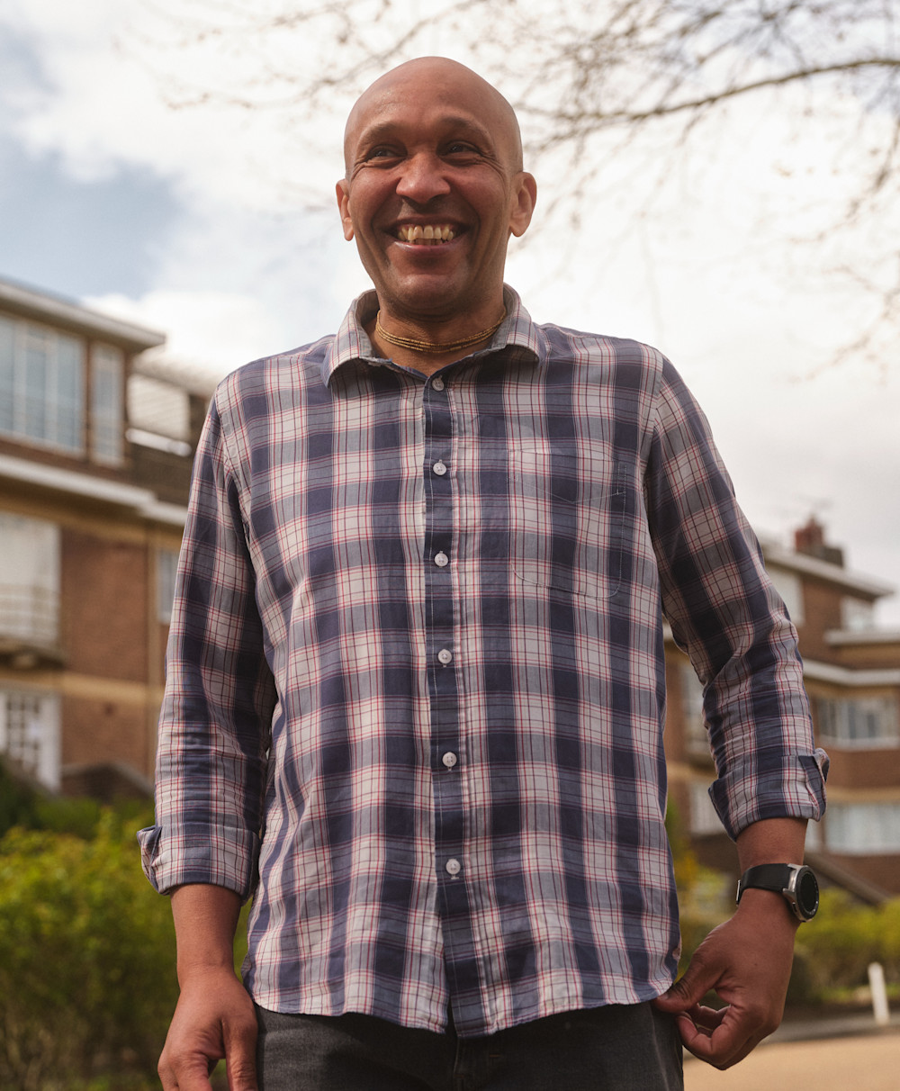 Saci smiles wearing a checked shirt outside in front of his home and a tree