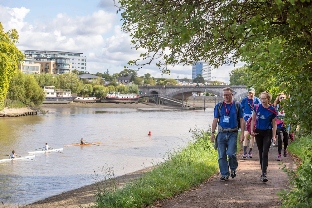 A group of four walkers hiking along the Thames riverside path. There are people rowing on the river and a view of London in the background.