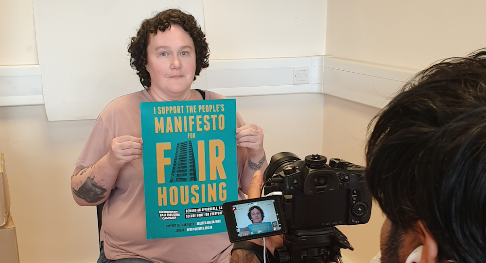 A seated campaigner holds the BFHC poster up to a video camera, from which someone is filming her.