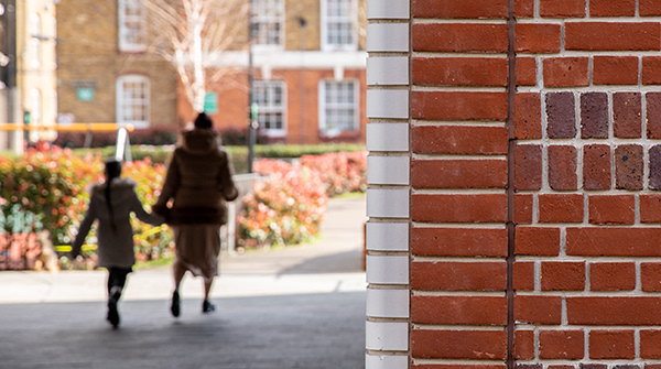 A mother and daughter are holding hands and walking through a brightly lit courtyard outside a block of flats.