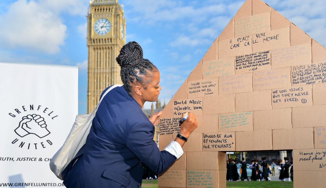 Wendy is writing on a wall of cardboard boxes at a campaign stunt. She stands beside a Grenfell United banner and Big Ben can be seen in the distance.