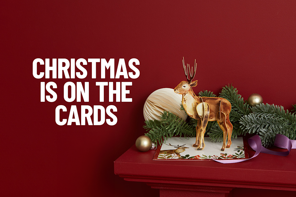 A festive red background with the text 'Christmas is on the cards'. A reindeer ornament stands on a Christmas card, surrounded by pine branches, gold ornaments, and a purple ribbon.