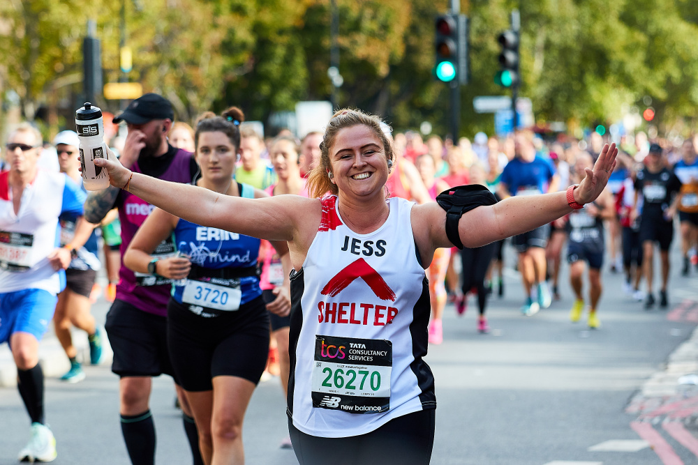 A woman with her arms outstretched running in the Marathon, wearing a Shelter jersey with the name 'Jess' printed on the front. 