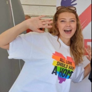 A picture of a woman with long, blonde hair and sunglasses. She is smiling and wearing a Shelter t-shirt which reads 'all love should have a home'. She is stood in front of a Shelter banner and has her arms raised excitedly.