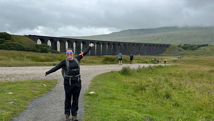 A woman wearing hiking gear holds her arms wide on a pathway, with an impressive view of a viaduct behind her.