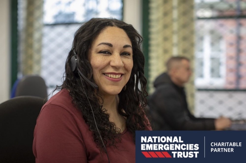 A smiling woman helpline advisor who is wearing headphones. In the corner is the National Emergencies Trust Charitable Partner logo.
