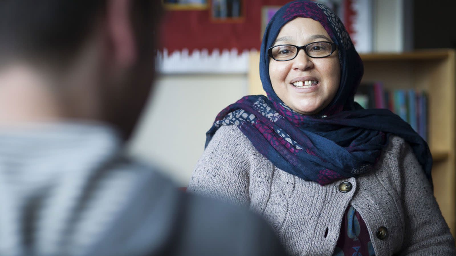 Woman in blue hijab and grey sweater talks with another person. Head scarf.