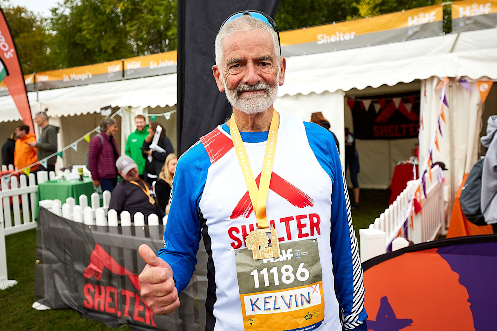 A Royal Parks Half Marathon runner wearing a Shelter vest and medal celebrates finishing his race. He has his thumbs up and is smiling at the camera.
