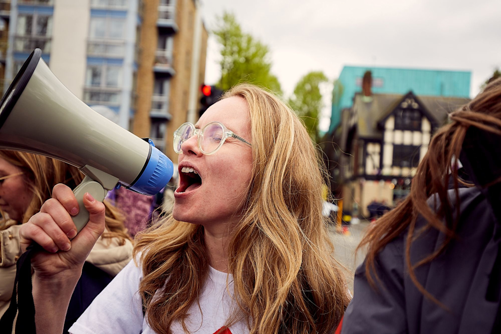 A woman wearing a Shelter top shouts into a megaphone, cheering on participants in a Shelter charity run.