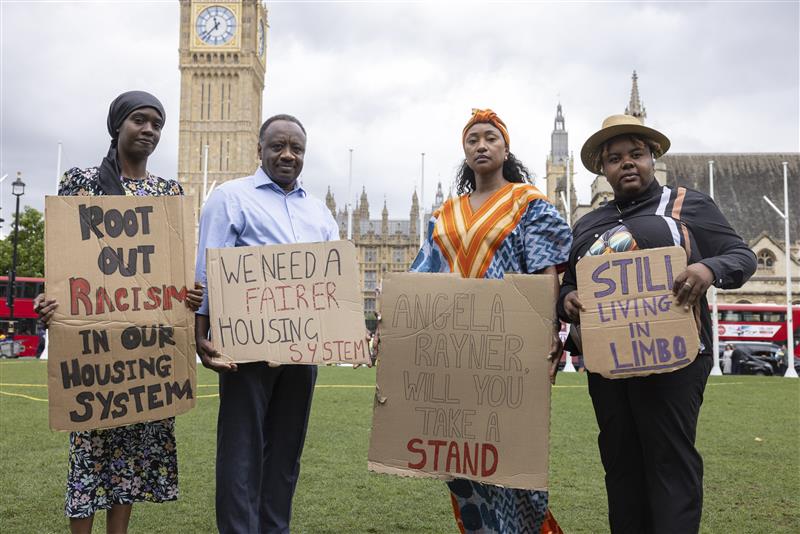 Four attendees from the Parliamentary launch event of the 'My colour speaks before me' racism and housing research stand on a lawn in front of Big Ben. They hold cardboard signs that talk about rooting out racism in the UK housing system.