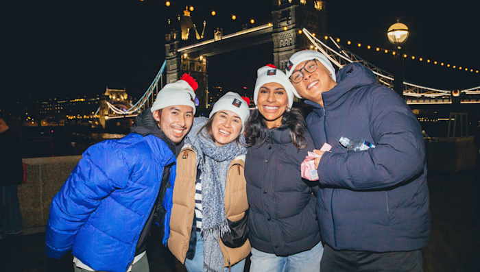 A group of four people wearing Walk for Home white and red bobble hats pose for a photo. It's night time and the lights of Tower Bridge are in the background.