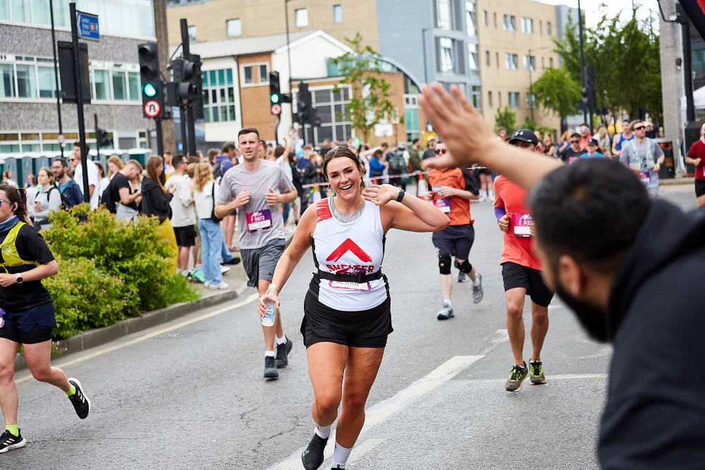 A woman runner wearing a Shelter vest approaches the Shelter cheer point, where an Events team member is holding out his hand for a high five.