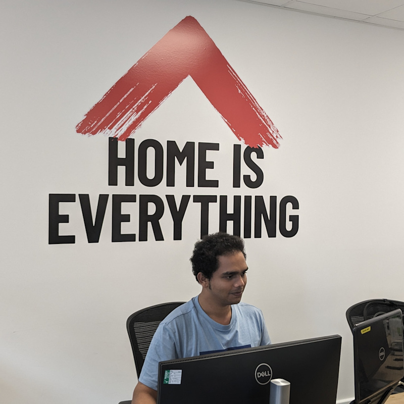 Saurabh is sat behind a desk at a computer. He has black curly hair and is wearing a blue t-shirt. Behind him is a white wall with the red roof logo of Shelter and Shelter Scotland. Beneath the roof is text reading: HOME IS EVERYTHING