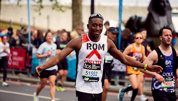 A man in a Shelter running vest passes the cheer point of a marathon, smiling and holding out his arms in celebration.