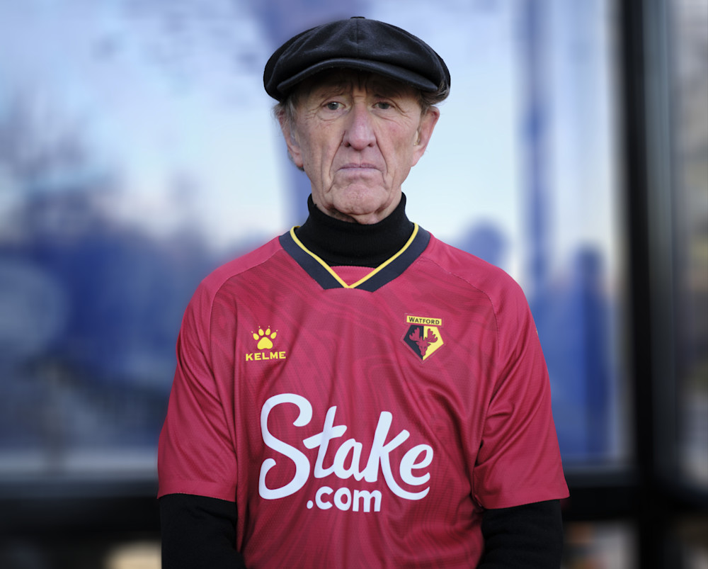 An older man in a cap in a red Watford away shirt