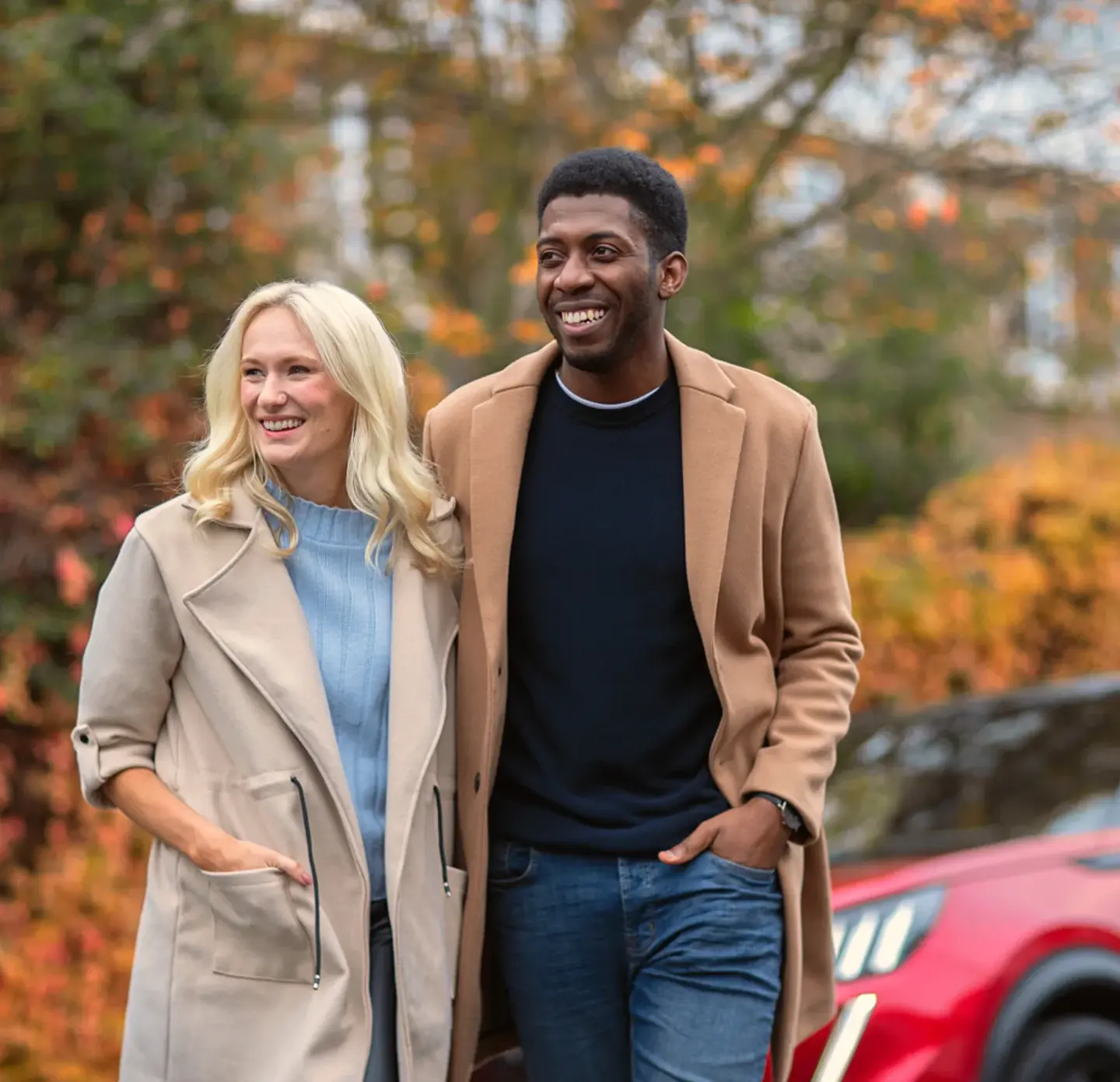 A couple with an arm around each other facing forward, looking away from the camera. Both are smiling and wearing brown coats, with a backdrop of autumnal leaves. Blurred in the background is the front of a red car.
