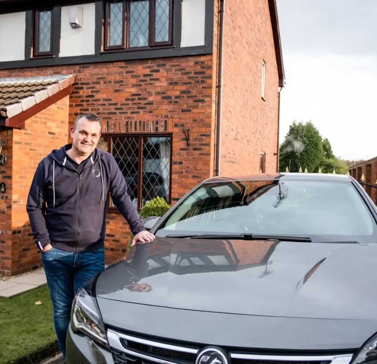 Barry standing, facing the camera and smiling, next to the front of his silver car. Pictured in the background is a house.