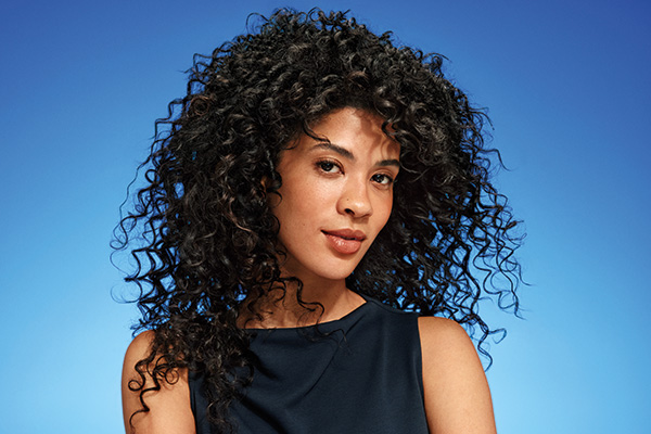 Donna con lunghi capelli neri naturalmente ricci su sfondo blu, che mette in risalto ricci definiti e capelli dall'aspetto sano.
