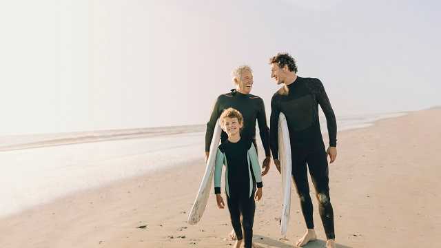 A grand-dad, dad and son walking on the beach in wet-suits with surfboards