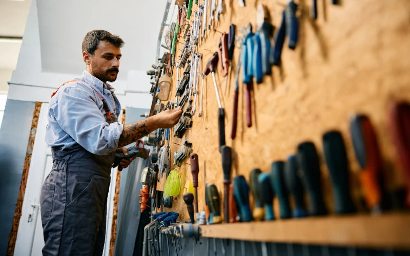 Ein Handwerker steht in einer Werkstatt vor einer Werkzeugwand und greift nach einem Werkzeug. Er trägt Arbeitskleidung und schaut konzentriert auf die Auswahl an Werkzeugen, die ordentlich an der Wand hängen.
