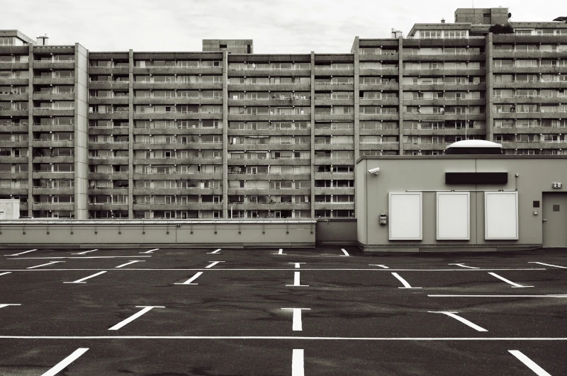 Ein sehr großer grauer, trister Wohnkomplex aus Beton vor einem leeren Parkplatz auf dem Dach einer grauen Parkgarage.
