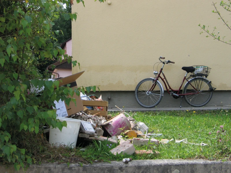 Fahrrad neben gelber Wand, Müllhaufen im Gras, Bäume und Sträucher im Hintergrund.