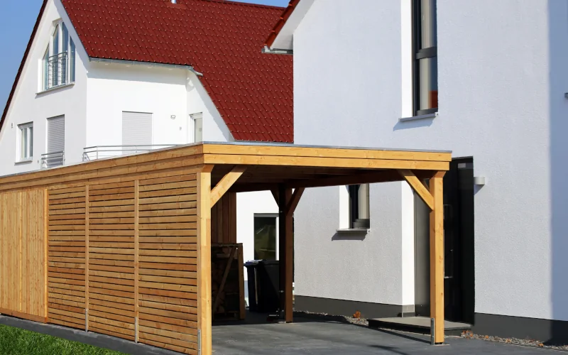 Holz Carport neben weißen Wohnhaus mit Mülltonnen darunter stehend