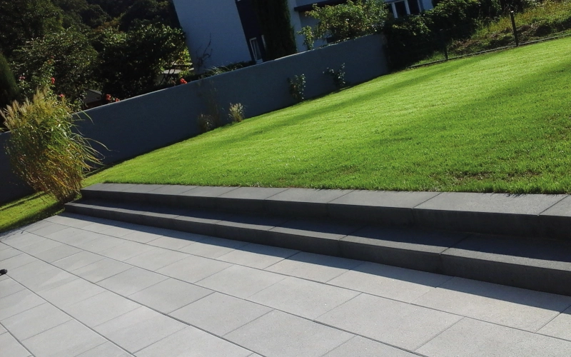 Moderne Terrasse mit hellgrauen Terrassenplatten und einem Stufensystem in anthrazit als Abgrenzung zu einer Rasenfläche.