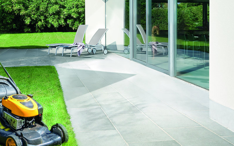 Terrasse mit grauen Fliesen neben einer Rasenfläche, auf der ein Rasenmäher steht