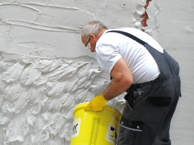 Ein älterer Mann hält einen gelben Eimer, dessen Masse er auf die Wand verteilt.