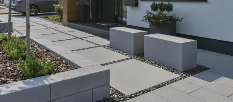 Eingangsbereich eines Hauses mit grauen Terassenplatten, ein wenig Kieselsteine dazwischen und einigen Pflanzen.