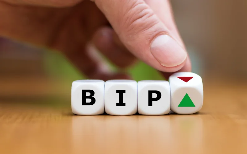 Vier Würfel nebeneinander, die in der Reihenfolge "BIP" stehen. Der letzte und vierte Würfel wird mit einer Hand so gekippt, dass man zwei Seiten des Würfels sieht. Auf der unteren Seite ist ein grünes Dreieck, auf der oberen ein rotes Dreieck.