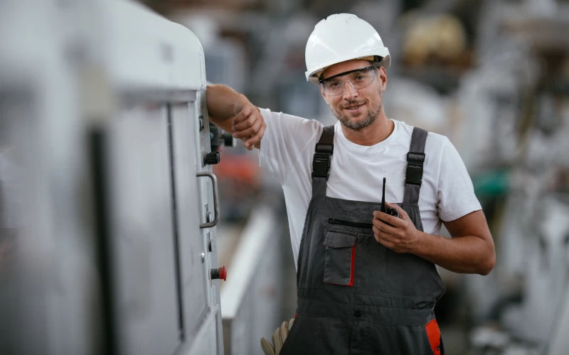 Auf dem Bild steht ein Mann mit Schutzhelm und Schutzbrille in einer Werkhalle. Er trägt Arbeitskleidung und hält ein Funkgerät in der Hand. Mit dem anderen Arm lehnt er sich locker an eine Maschine und blickt lächelnd in die Kamera.