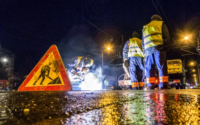 Bauarbeiter arbeiten Nachts an einer Straße, im Vordergrund ein Baustellenschild